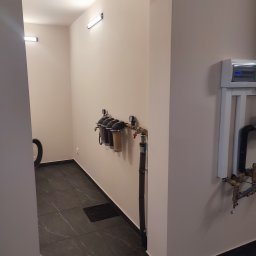 Knopik Rafał Lipusz - Wąskie pomieszczenie gospodarcze z zamontowanym systemem filtracji wody i rurami na beżowej ścianie, obok skrzynka elektryczna. Ciemne płytki podłogowe, nowoczesne oświetlenie LED.