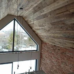 Poddasze z drewnianą boazerią na skośnym suficie, duże trójkątne okno z widokiem na zimowy krajobraz, fragment ściany z czerwonej cegły i wiszące lampy bez kloszy.