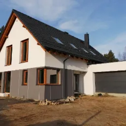 Nowoczesny dom jednorodzinny z dwukolorową elewacją, ciemną dachówką i przylegającym garażem, w trakcie prac wykończeniowych na działce.