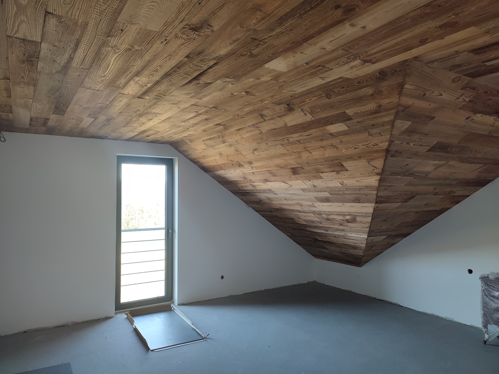 Wykończone drewnem poddasze ze skośnym sufitem i oknem. Widoczne detale drewnianej konstrukcji sufitu i surowa podłoga. Charakterystyczny kształt poddasza.