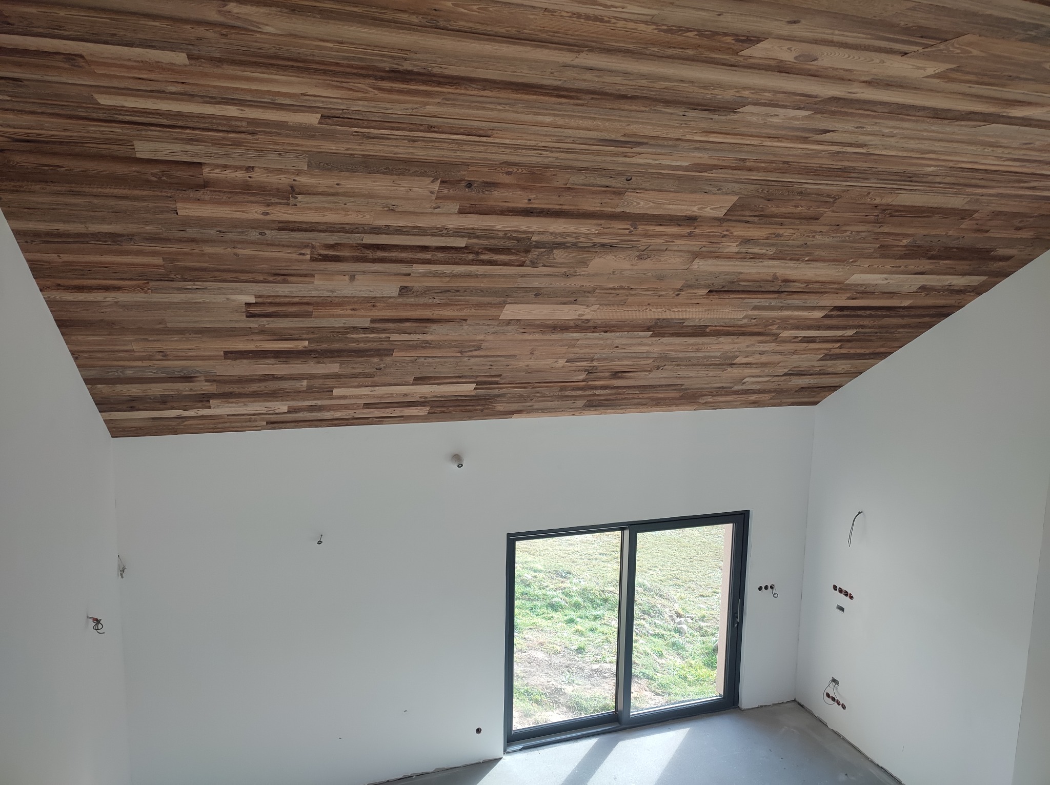 Drewniana boazeria na skośnym suficie w surowym wnętrzu z oknem. Widoczne wystające kable elektryczne. Podłoga betonowa. Naturalne światło dzienne.