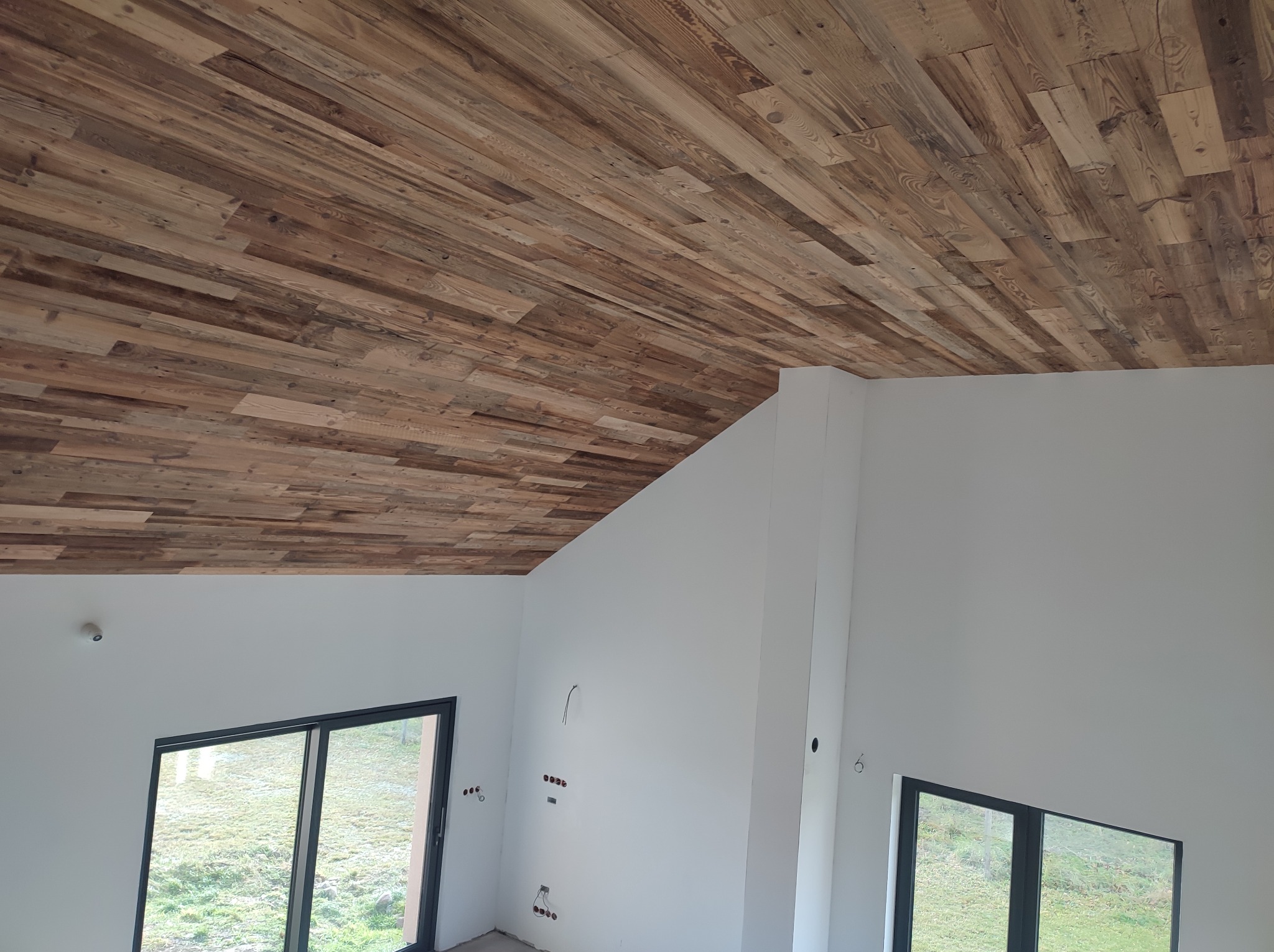 Drewniana boazeria na skośnym suficie w jasnym wnętrzu z oknami. Widoczne kable elektryczne na ścianach, co sugeruje etap wykończeniowy. Nietypowy układ desek.