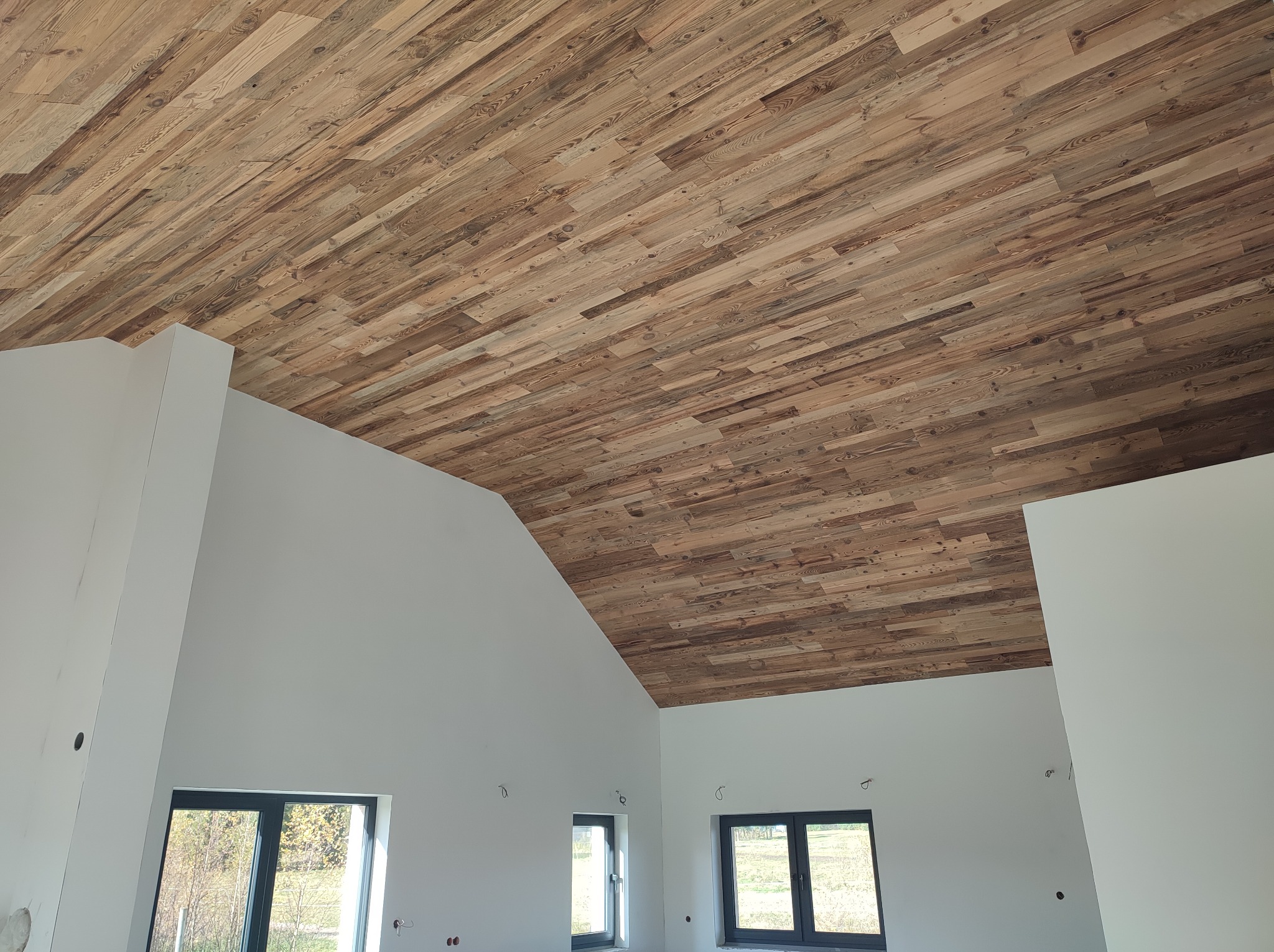 Drewniany sufit z desek w jasnym wnętrzu z oknami. Deski ułożone pod kątem, tworząc unikalny wzór. Ściany białe, surowe wykończenie.