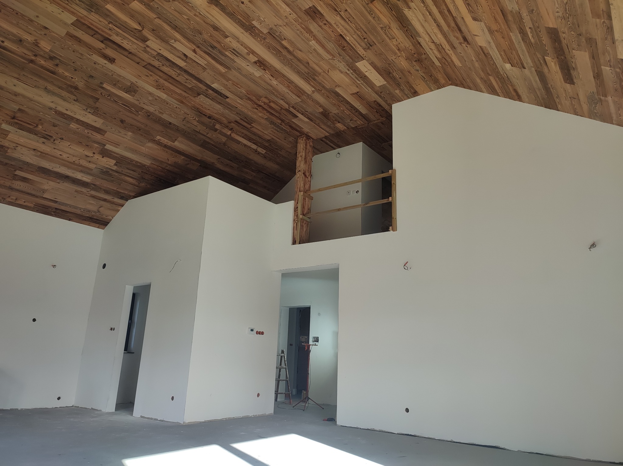 Surowe wnętrze z drewnianym sufitem z desek, białe ściany, otwory okienne i drzwiowe. Widoczna drewniana kolumna i fragment balustrady. Podłoga betonowa, wpadające światło.