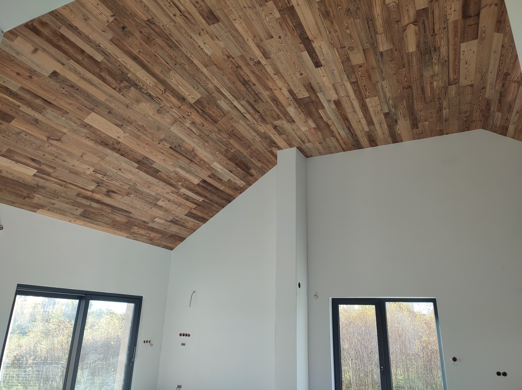 Drewniana boazeria na skośnym suficie w nowym domu z oknami. Naturalne drewno tworzy ciepły kontrast z białymi ścianami. Widoczne przygotowania do instalacji elektrycznej.