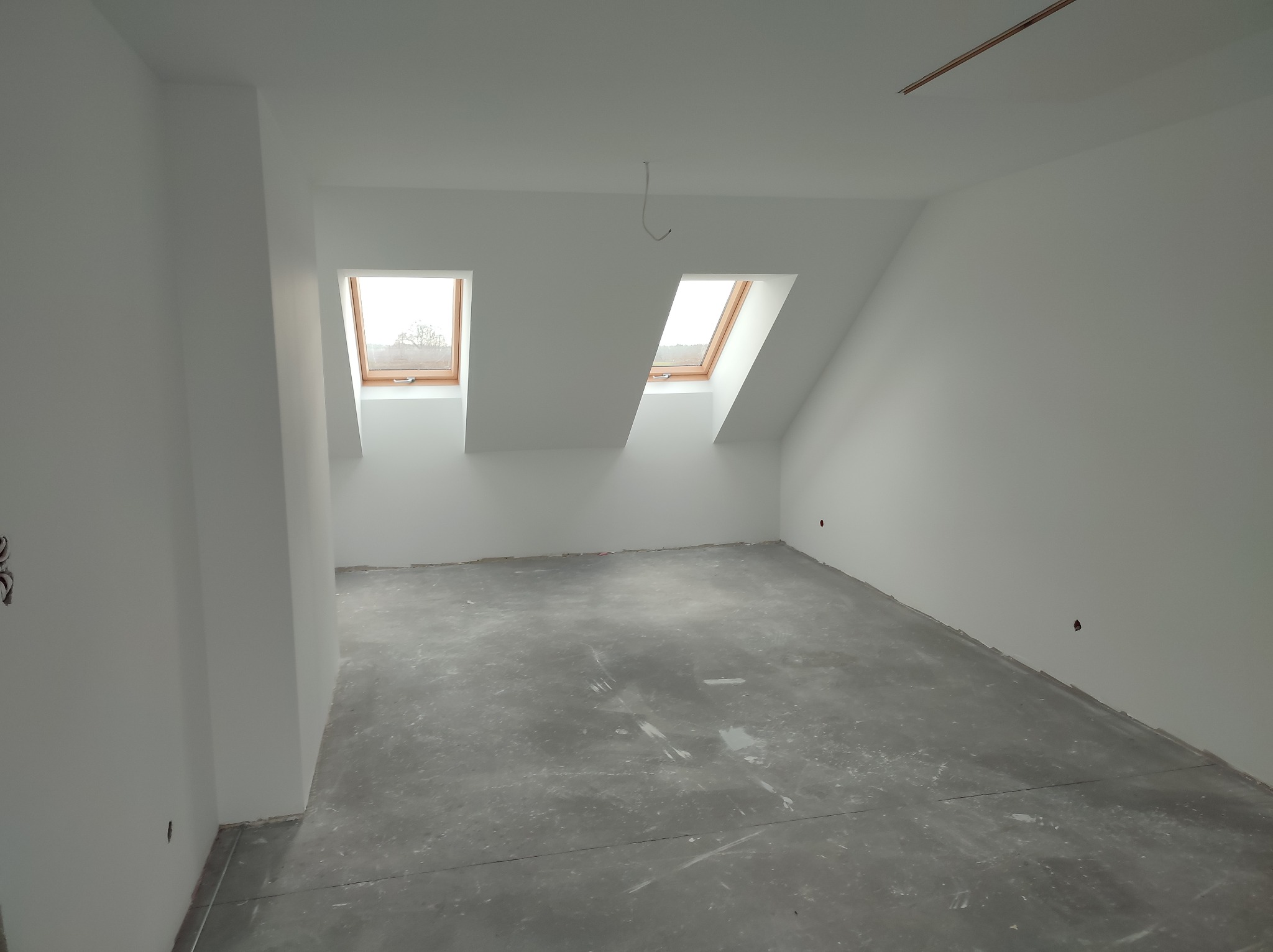 Poddasze po remoncie: białe ściany, dwa okna dachowe z drewnianą ramą, szara podłoga w stanie surowym, widoczne przewody elektryczne. Minimalistyczne wykończenie.