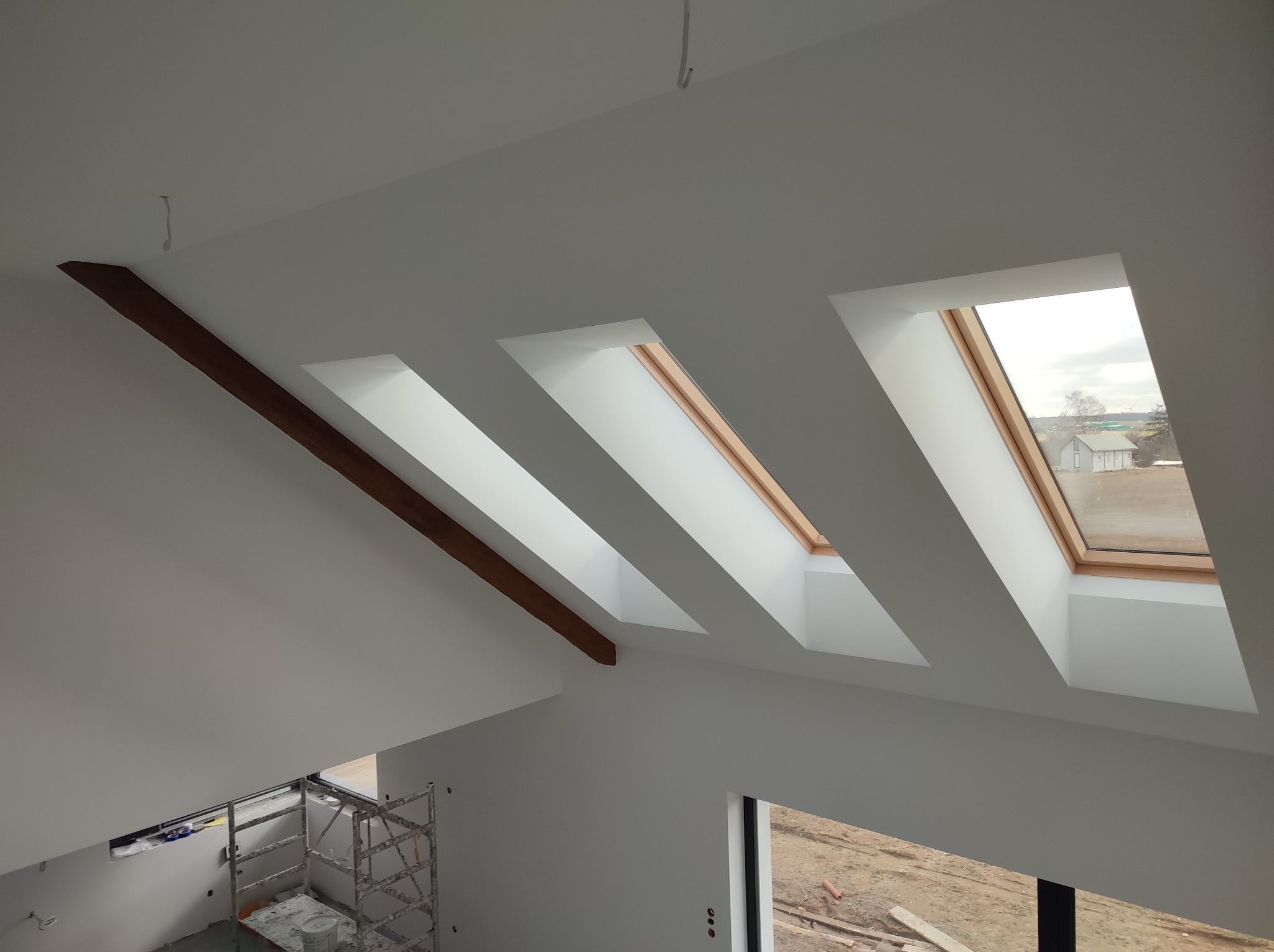 Białe poddasze z trzema oknami dachowymi i drewnianą belką. Widoczne rusztowanie i przygotowane ściany do dalszych prac wykończeniowych.