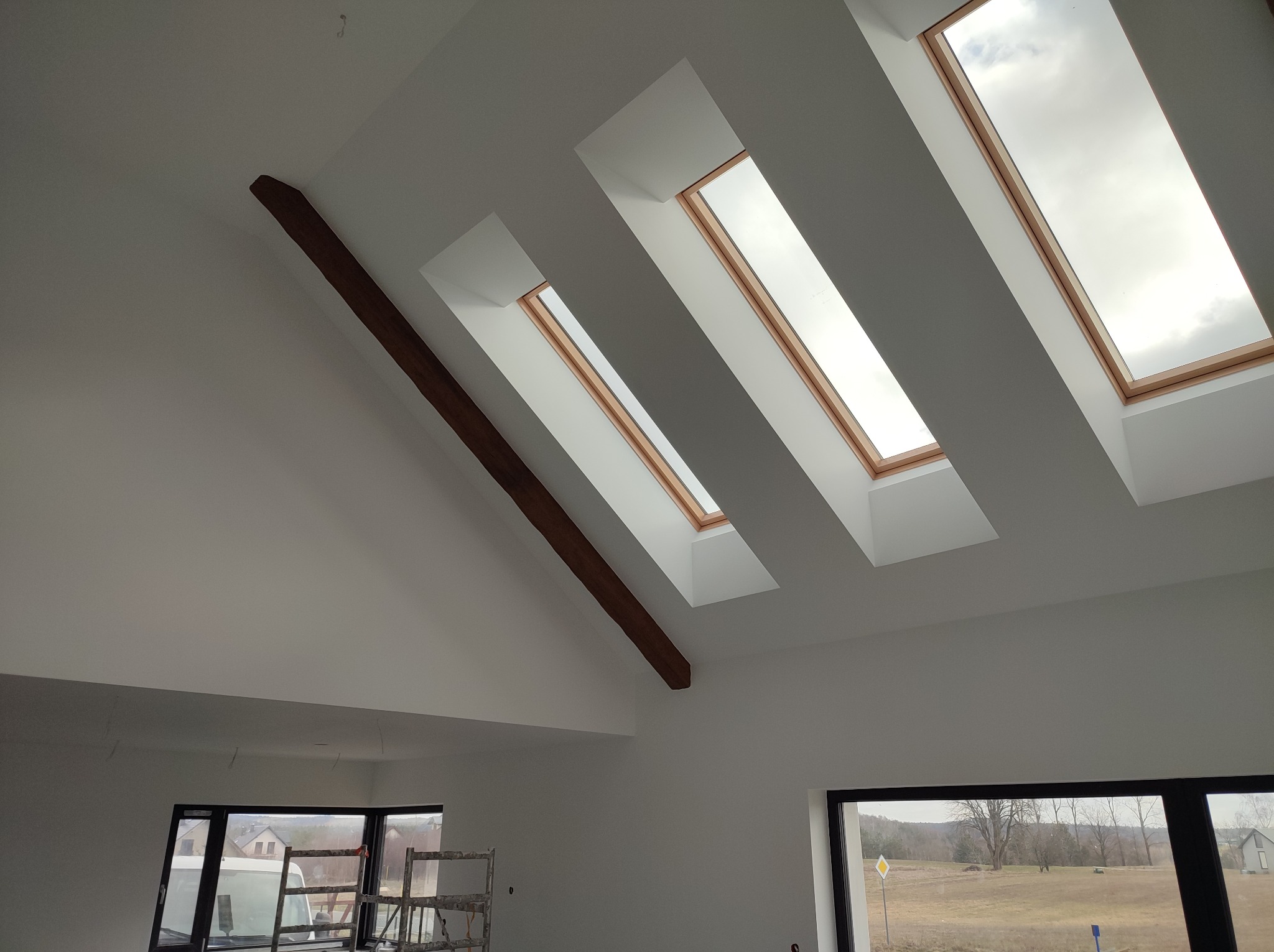 Poddasze po malowaniu na biało z trzema oknami dachowymi i drewnianą belką stropową. Widoczne rusztowania i okno z widokiem na zewnątrz.