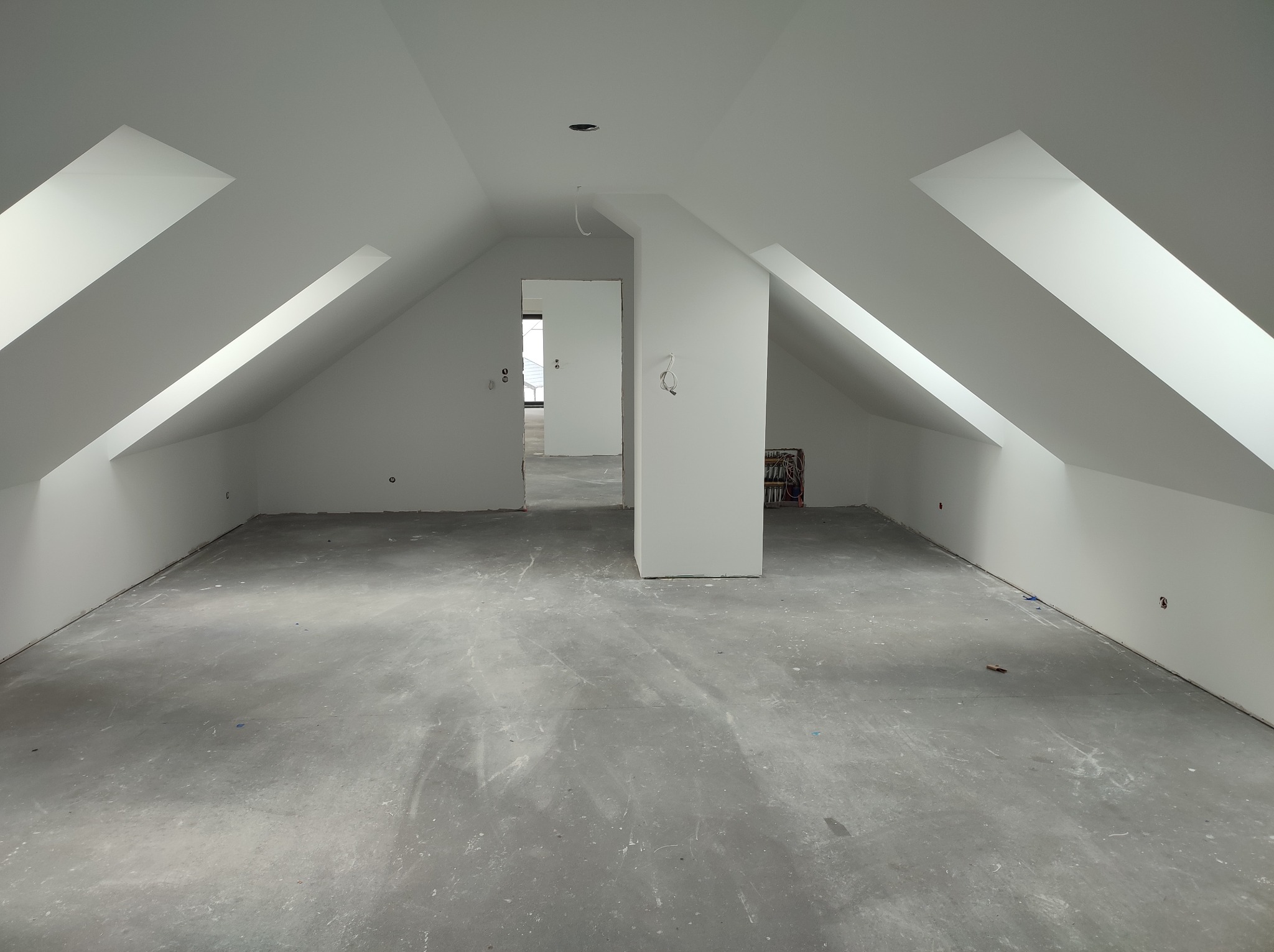 Poddasze po remoncie, świeżo pomalowane na biało, z widocznymi skosami i oknami dachowymi. Surowa podłoga, przygotowana do dalszych prac wykończeniowych. Widok na kolejne pomieszczenia.