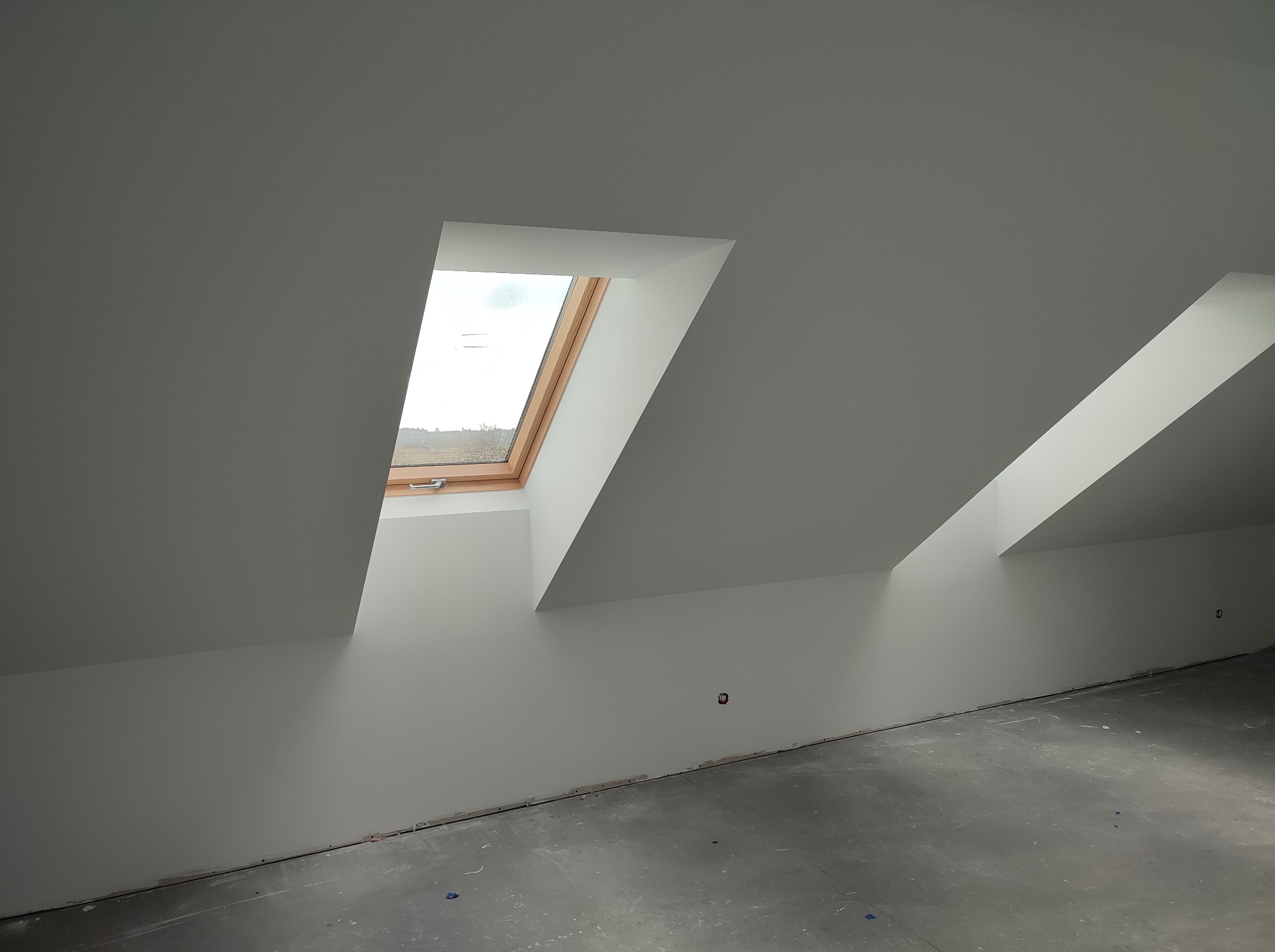 Poddasze po remoncie: białe ściany, okno dachowe z drewnianą ramą, szara podłoga. Widoczne ślady po pracach wykończeniowych przy podłodze. Minimalistyczny styl.