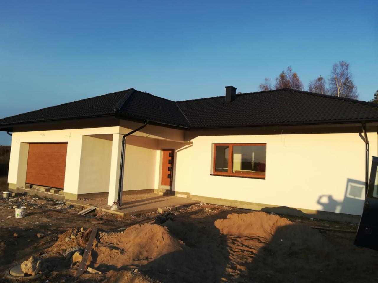 Nowo wybudowany dom jednorodzinny z garażem, z ciemną dachówką i jasną elewacją, w trakcie prac wykończeniowych na zewnątrz, widoczne pryzmy piasku.