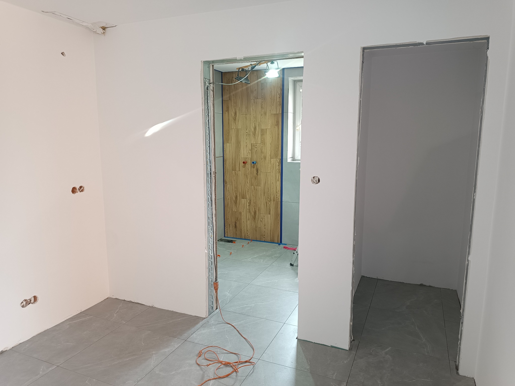 Wnętrze w trakcie remontu w Lipuszu: widoczne przygotowane ściany do malowania, ułożona podłoga z szarych płytek oraz fragment wykończonej łazienki z drewnopodobnymi panelami na ścianie.