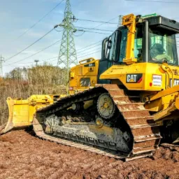 Żółty buldożer marki CAT na nierównym, gliniastym terenie, z widocznymi śladami gąsienic, w tle słupy wysokiego napięcia i drzewa bez liści.