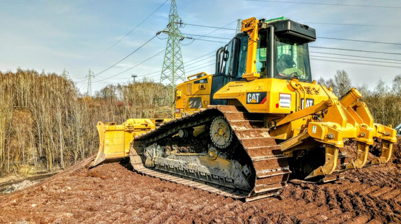 Żółty buldożer marki CAT na nierównym, gliniastym terenie, z widocznymi śladami gąsienic, w tle słupy wysokiego napięcia i drzewa bez liści.