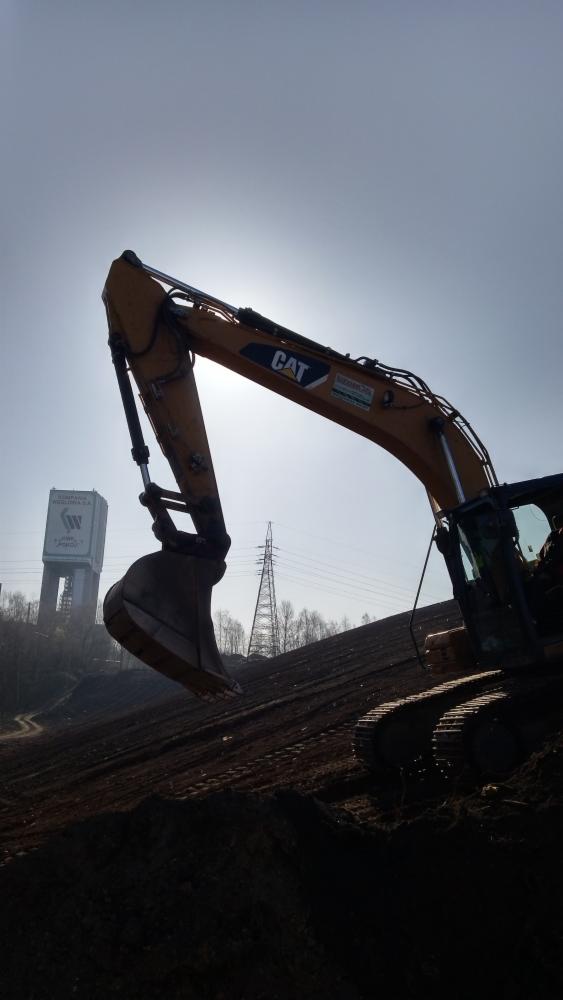 Żółta koparka CAT na tle nasypu ziemnego, w tle wieża ciśnień i słup wysokiego napięcia, ujęcie pod słońce.