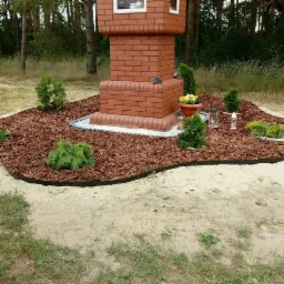 Aranżacja wokół murowanej kapliczki: młode drzewka iglaste, kwiaty w doniczce i znicze, kora rozsypana na rabacie z obrzeżem, całość na piaszczystym podłożu.