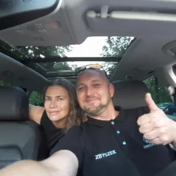 Uśmiechnięty kierowca z pasażerką w samochodzie z panoramicznym dachem, kierowca pokazuje kciuk w górę, na koszulce widoczne logo firmy transportowej.