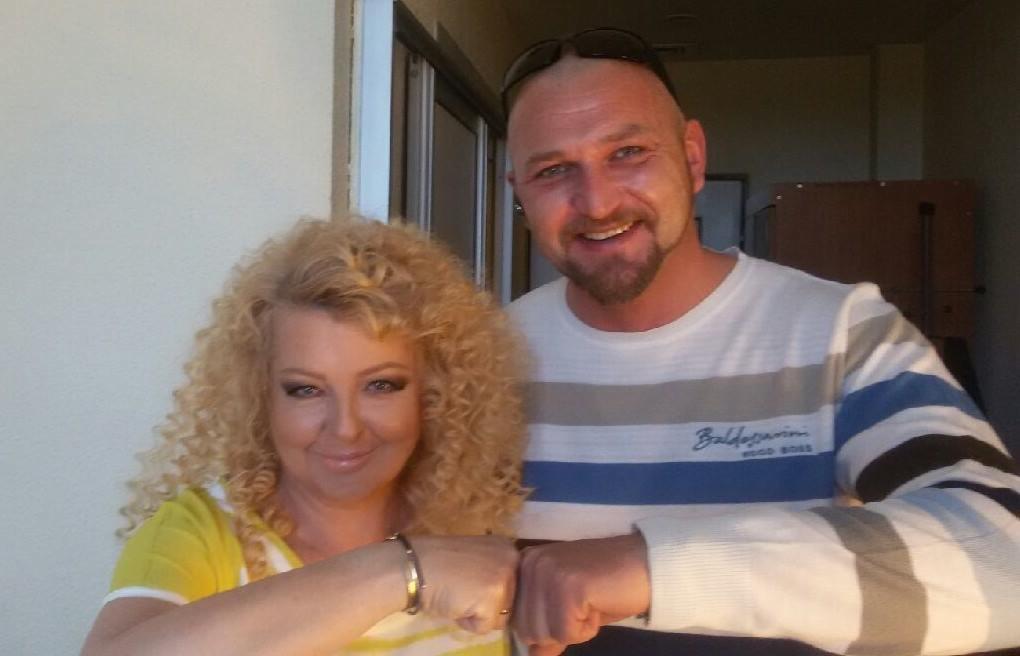 Uśmiechnięta kobieta z burzą blond loków i mężczyzna z brodą, ubrany w koszulkę w paski, witają się 'żółwikiem' na tle jasnej ściany i drzwi.