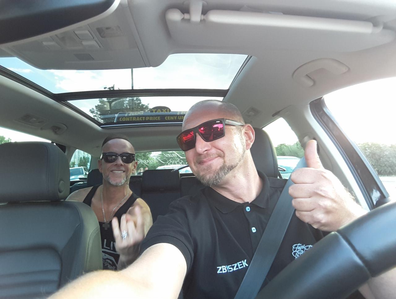 Kierowca z pasażerem w samochodzie z panoramicznym dachem, kierowca w koszulce polo z logo firmy, kciuk w górę.