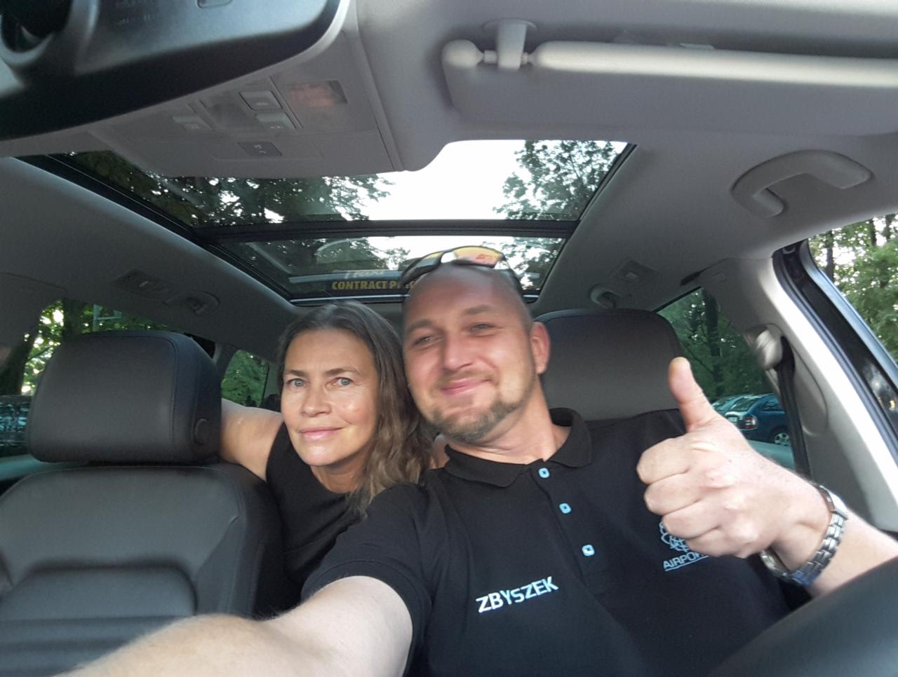 Uśmiechnięty kierowca z pasażerką w samochodzie z panoramicznym dachem, kierowca pokazuje kciuk w górę, na koszulce widoczne logo firmy transportowej.