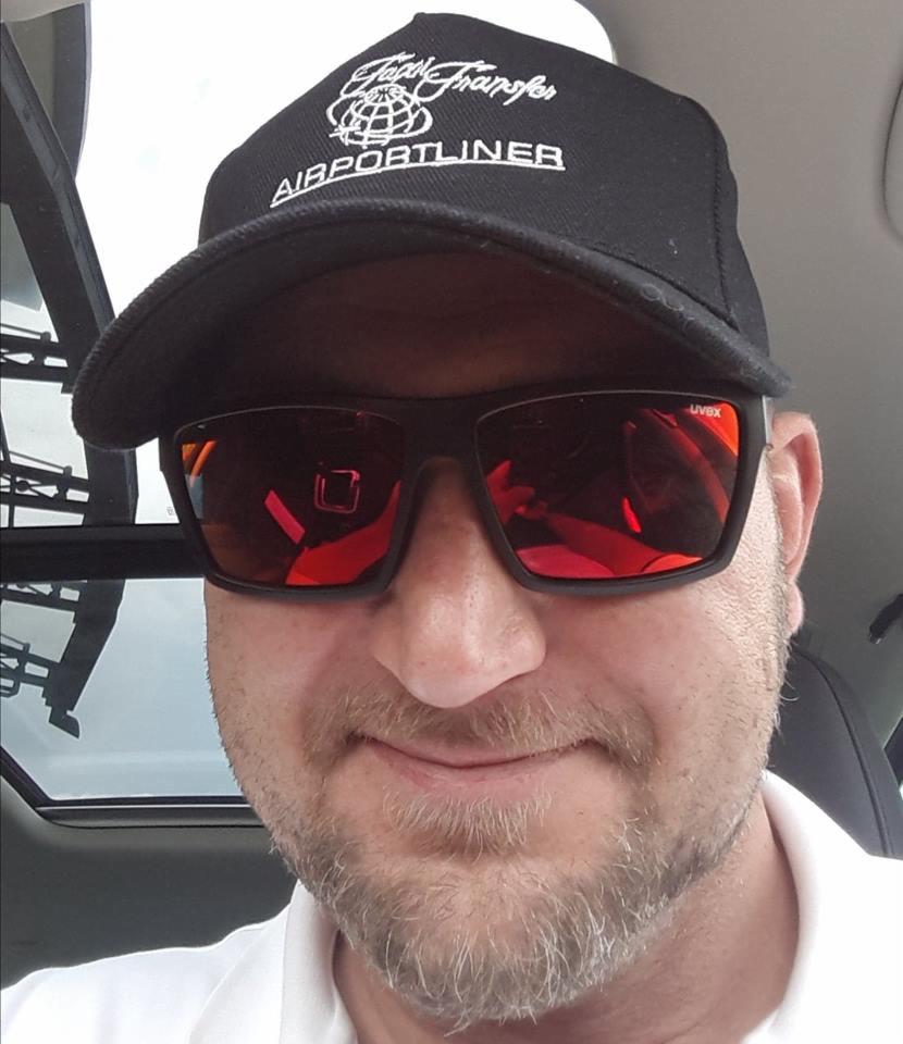 Portret mężczyzny w czapce z napisem 'Airportliner' i okularach przeciwsłonecznych Uvex, siedzącego w samochodzie z widocznym dachem panoramicznym.