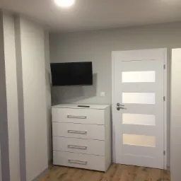 Szare ściany z pionowymi pasami w nowym pokoju, biała komoda z telewizorem na ścianie, białe drzwi z przeszkleniami i szafa wnękowa, drewnopodobna podłoga.