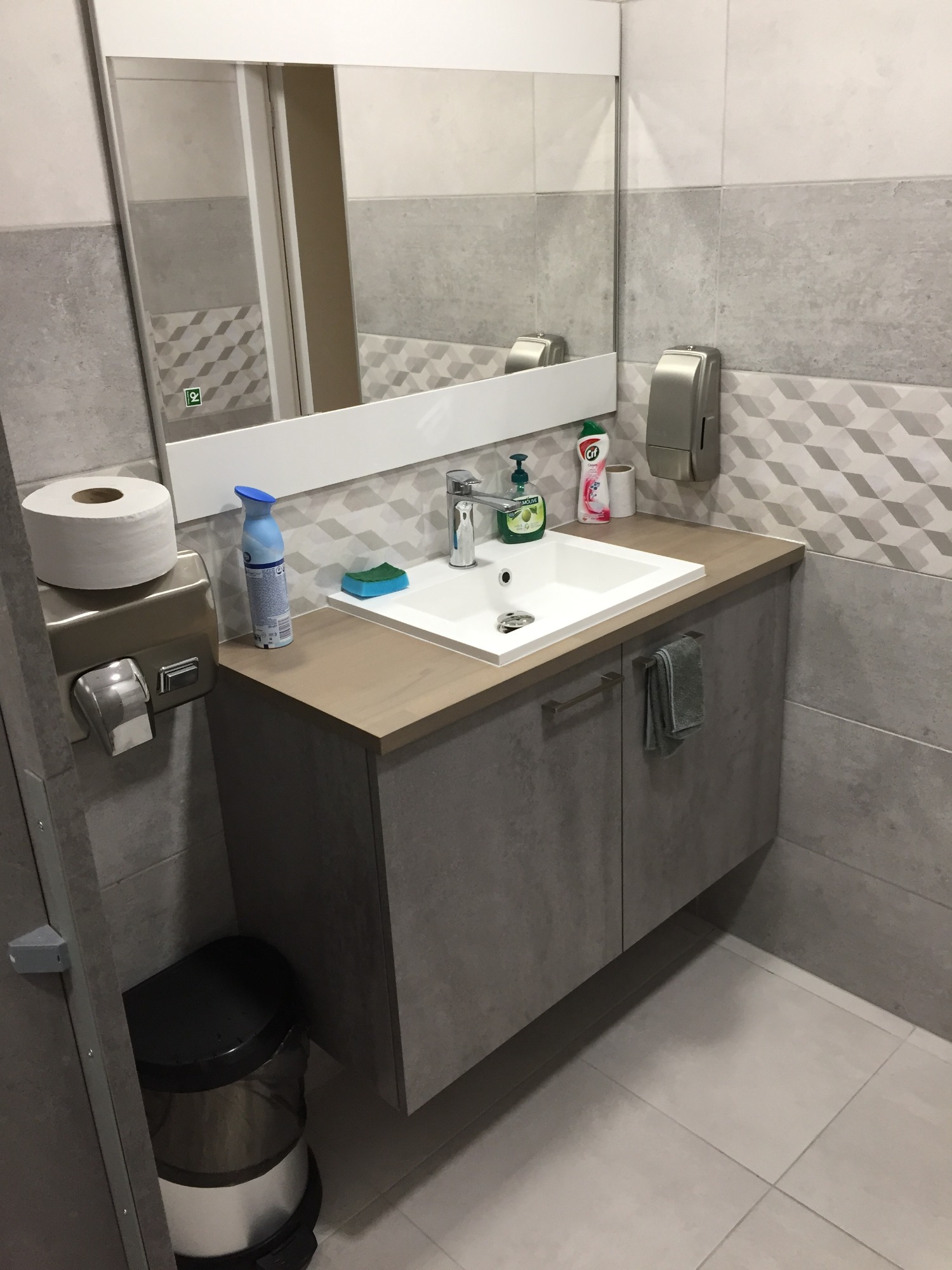 Szaro-biała łazienka z umywalką nablatową, baterią, dozownikiem mydła, ręcznikiem, rolką papieru toaletowego i odświeżaczem powietrza na blacie. W tle lustro i wzorzyste płytki.