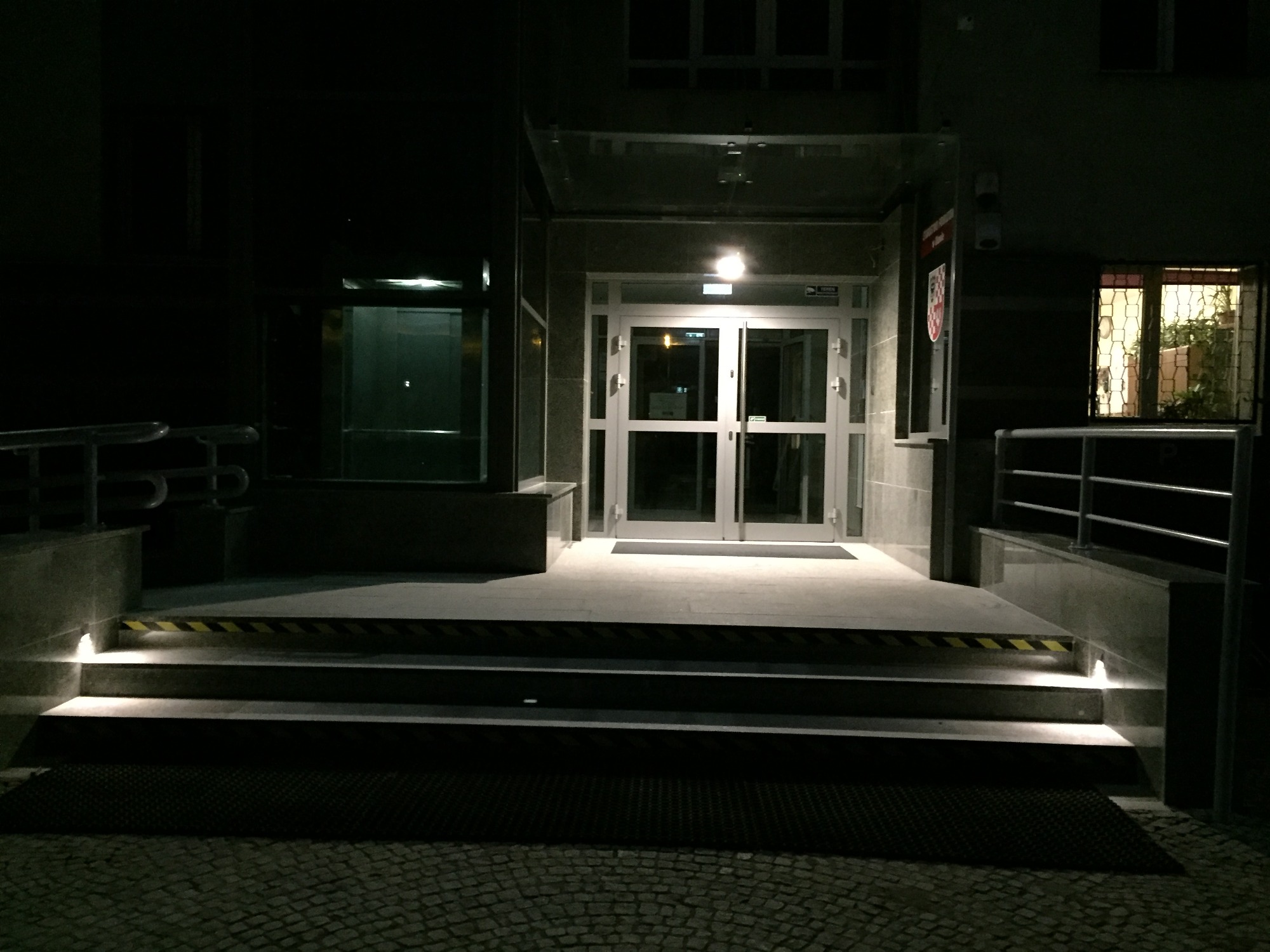 Nocne ujęcie wejścia do Starostwa Powiatowego w Oławie: szklane drzwi wejściowe z oświetleniem, schody z pasami ostrzegawczymi, elewacja budynku.