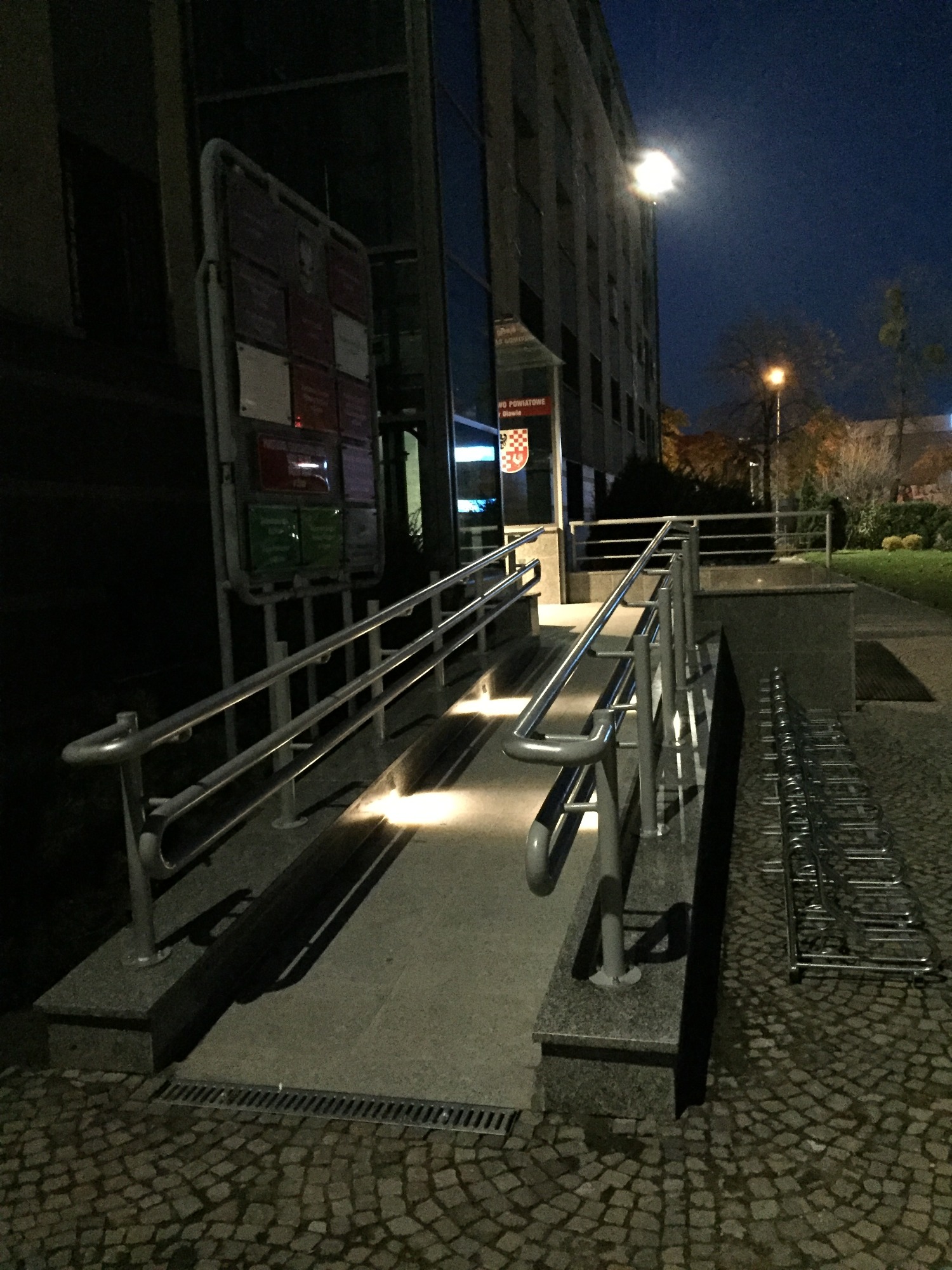 Zewnętrzny podjazd dla niepełnosprawnych z metalowymi barierkami, oświetlony punktowo, przed budynkiem Starostwa Powiatowego w Oławie, widok nocny.