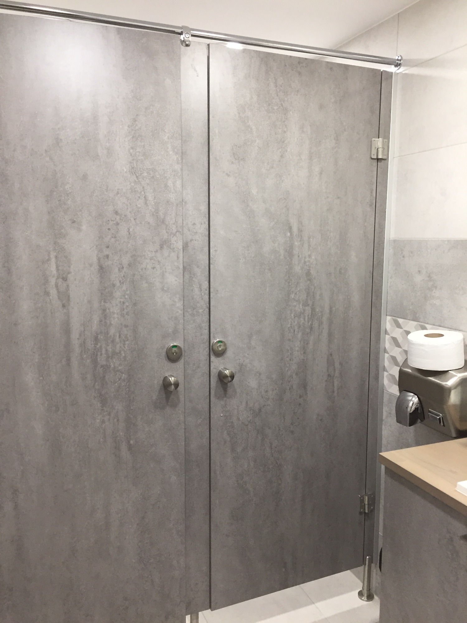 Szare kabiny toaletowe z metalowymi zamkami i systemem przesuwnym, w tle dozownik papieru i suszarka do rąk.