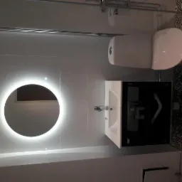 Nowoczesna łazienka z okrągłym lustrem LED, białą umywalką z czarną szafką, toaletą i wzorzystymi płytkami podłogowymi.