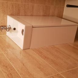 AGATE Sp. z o.o. - Kompaktowa toaleta z podtynkową spłuczką i chromowaną armaturą, zamontowana w narożniku wyłożonym beżowymi płytkami imitującymi trawertyn.