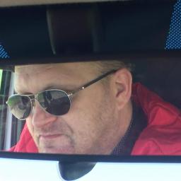 AGATE Sp. z o.o. - Mężczyzna w okularach przeciwsłonecznych, widziany w lusterku wstecznym samochodu. Czerwona kurtka, wzorzysta koszula, krajobraz za oknem.