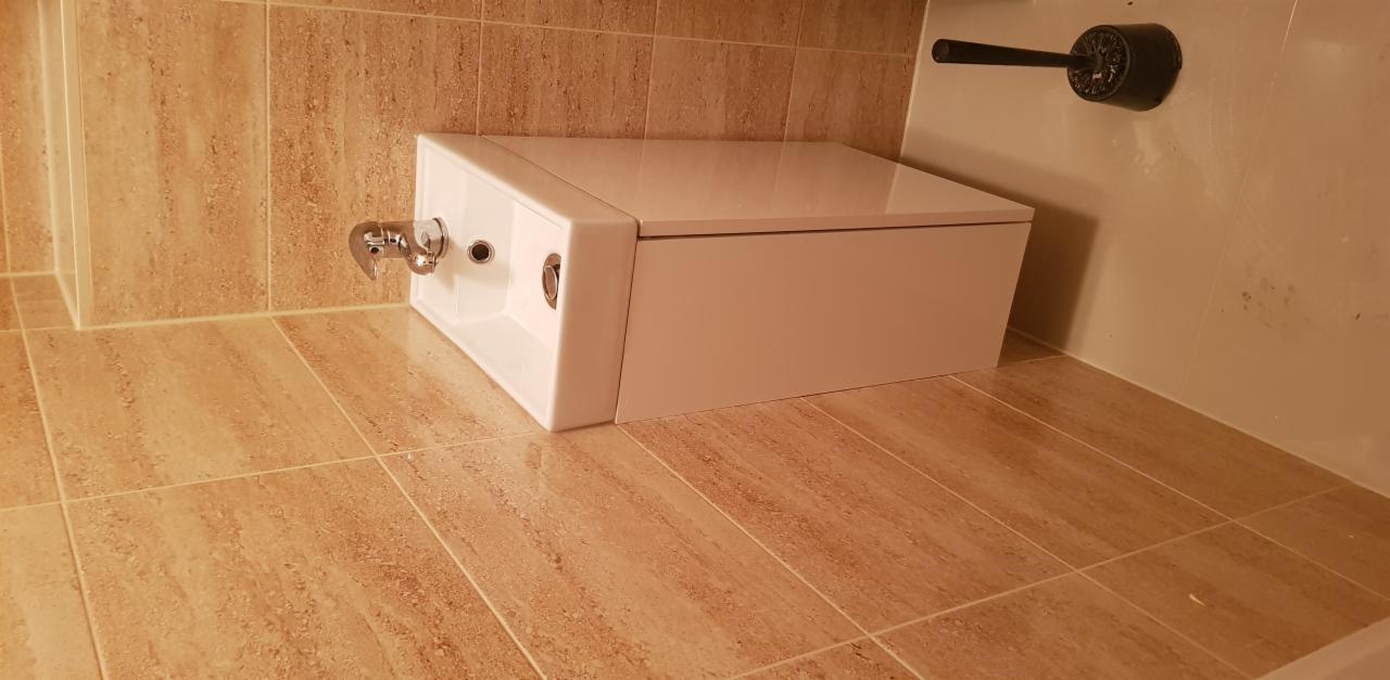 Kompaktowa toaleta z podtynkową spłuczką i chromowaną armaturą, zamontowana w narożniku wyłożonym beżowymi płytkami imitującymi trawertyn.