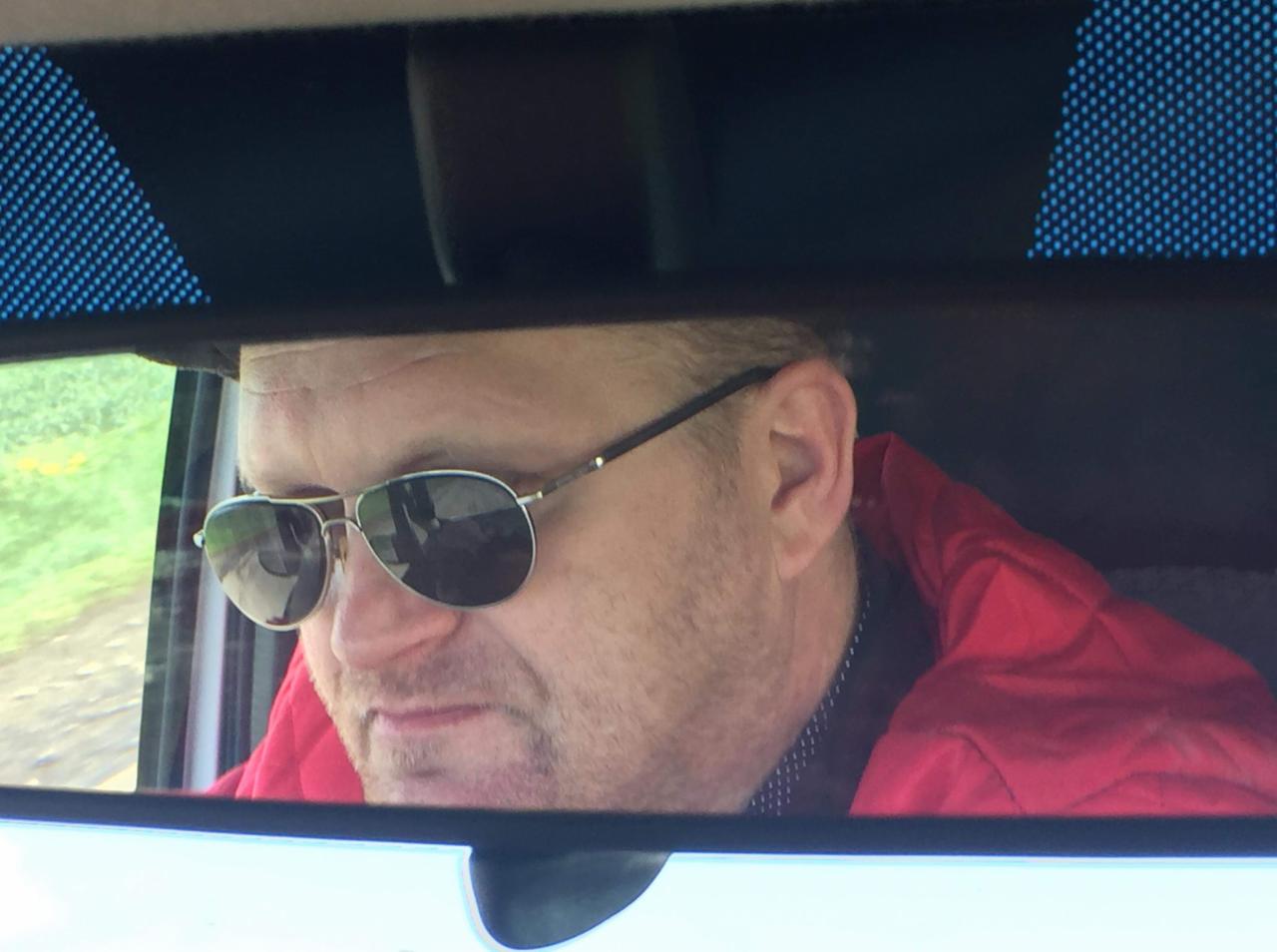 Mężczyzna w okularach przeciwsłonecznych, widziany w lusterku wstecznym samochodu. Czerwona kurtka, wzorzysta koszula, krajobraz za oknem.