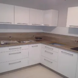 Biała, narożna zabudowa kuchenna z drewnianym blatem i frontami z podłużnymi, metalowymi uchwytami, widoczne przygotowane otwory na gniazdka elektryczne.