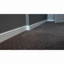 Ciemnobrązowa podłoga pokryta wykładziną dywanową w drobne, żółte kropeczki, z białymi listwami przypodłogowymi na tle szarej ściany i fragmentu drzwi.