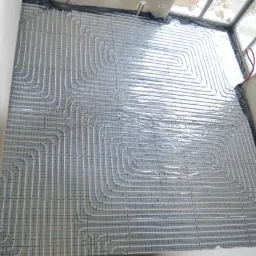 Białe rurki ogrzewania podłogowego ułożone w pętle na folii z siatką, widoczne fragmenty ścian i okna w surowym stanie.