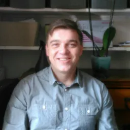 Portret uśmiechniętego mężczyzny w szarej koszuli na tle regału z pudełkami i rośliną doniczkową