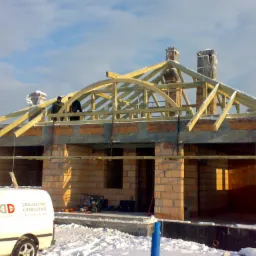 Budowa domu w zimowej scenerii: murowane ściany, drewniana konstrukcja więźby dachowej z łukowym elementem, kominy zabezpieczone folią, śnieg na dachu i wokół budynku, firmowy samochód z napisem...