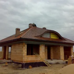 Budowa domu jednorodzinnego w trakcie prac dekarskich, widoczni dekarze na dachu z cegły ceramicznej Roben Poroton, ściany z pustaków ceramicznych, drabina oparta o ścianę, pochmurne niebo w tle.