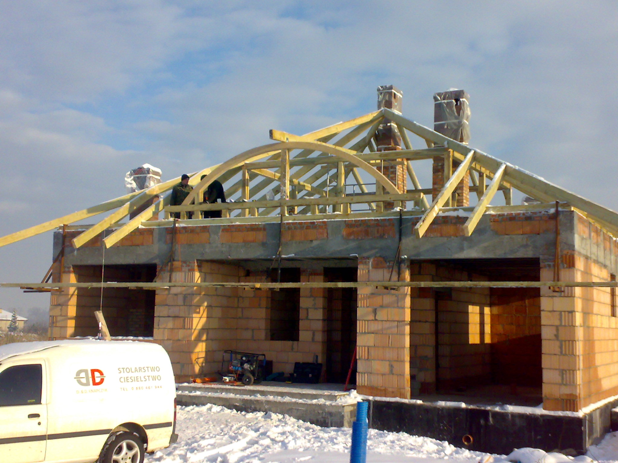 Budowa domu w zimowej scenerii: murowane ściany, drewniana konstrukcja więźby dachowej z łukowym elementem, kominy zabezpieczone folią, śnieg na dachu i wokół budynku, firmowy samochód z napisem...