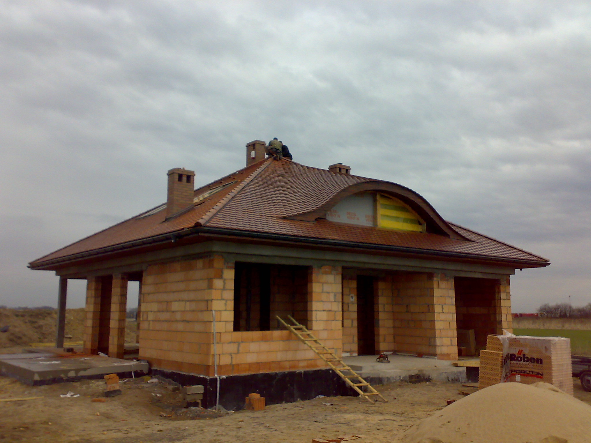 Budowa domu jednorodzinnego w trakcie prac dekarskich, widoczni dekarze na dachu z cegły ceramicznej Roben Poroton, ściany z pustaków ceramicznych, drabina oparta o ścianę, pochmurne niebo w tle.