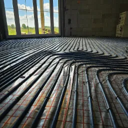 Rozłożone rury ogrzewania podłogowego na folii z czerwoną siatką, widok z niskiej perspektywy w surowym wnętrzu z dużym oknem, przez które widać błękitne niebo.