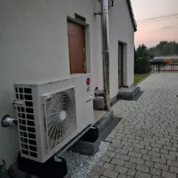 Biała jednostka zewnętrzna z logo LG, zamontowana na betonowych bloczkach i metalowej konstrukcji przy elewacji domu z brązowymi drzwiami i kominem ze stali nierdzewnej, na tle kostki brukowej...
