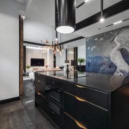 Vanilla Design - Czarne szafki kuchenne z dekoracyjnymi, złotymi wykończeniami, na tle salonu z tapetą przedstawiającą czaplę i nowoczesnym oświetleniem.