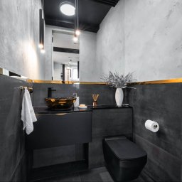 Vanilla Design - Nowoczesna łazienka w odcieniach szarości i czerni z akcentami złota, umywalka w kolorze starego złota, minimalistyczna toaleta, dekoracyjny wazon z suchymi gałązkami, lustro z oświetleniem.