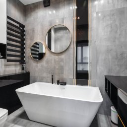 Vanilla Design - Nowoczesna łazienka z wolnostojącą białą wanną, szarą płytką imitującą beton, okrągłymi lustrami w złotych ramach i czarnymi akcentami, takimi jak grzejnik drabinkowy i blat umywalkowy.