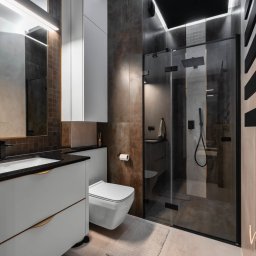 Vanilla Design - Nowoczesna łazienka w odcieniach brązu i szarości, z kabiną prysznicową typu walk-in, podwieszaną toaletą i umywalką na szafce z czarnym blatem, oświetlona listwą LED nad lustrem.
