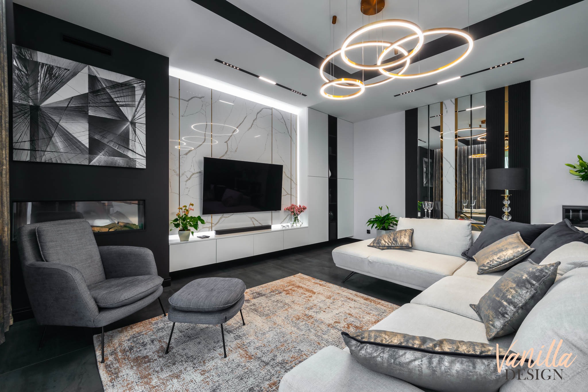 Nowoczesny salon z szarą sofą narożną, fotelem z podnóżkiem, marmurową ścianą za telewizorem i oryginalnym oświetleniem pierścieniowym.